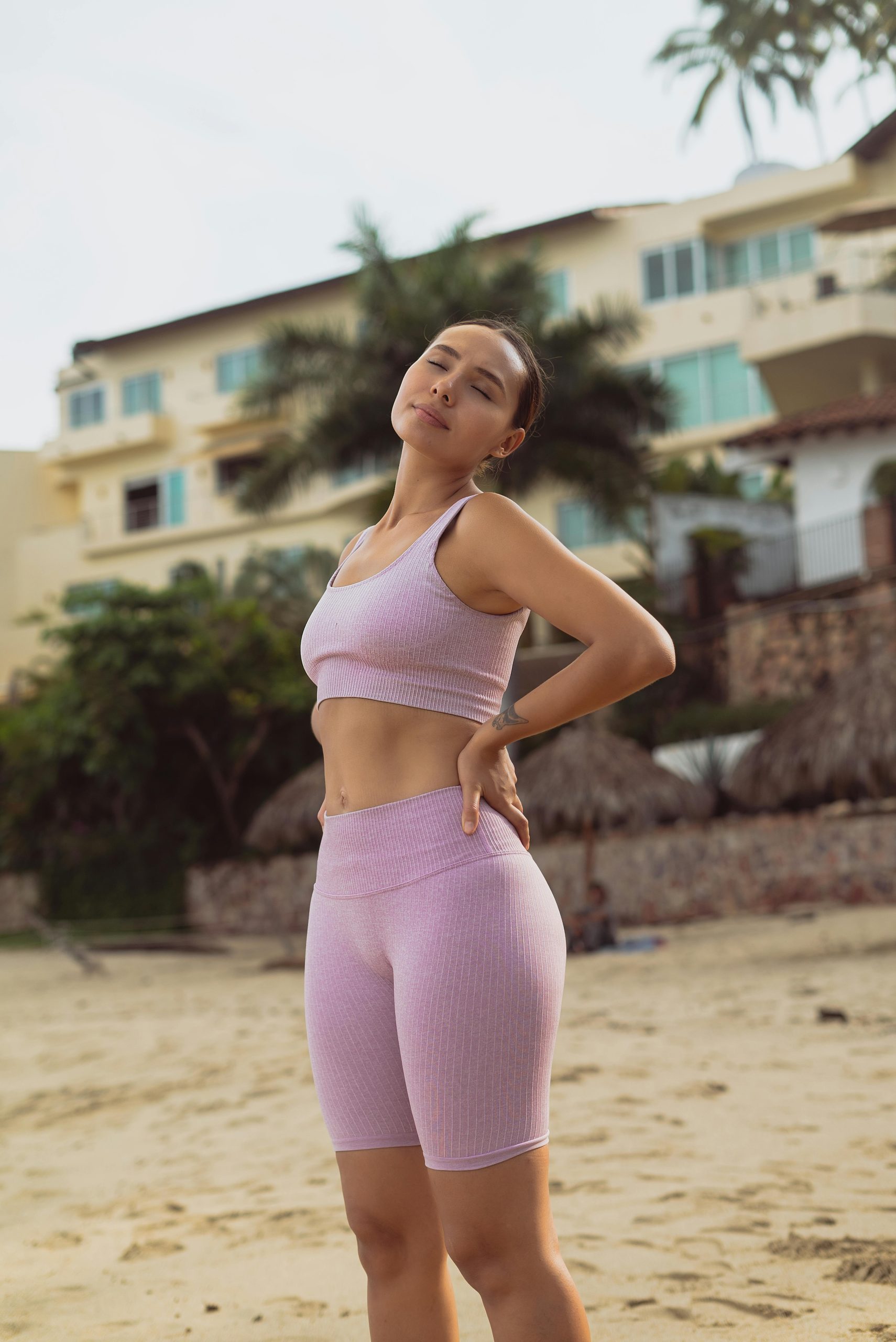 Fit woman in sports clothing stretching on a beach with palm trees and buildings in the background.