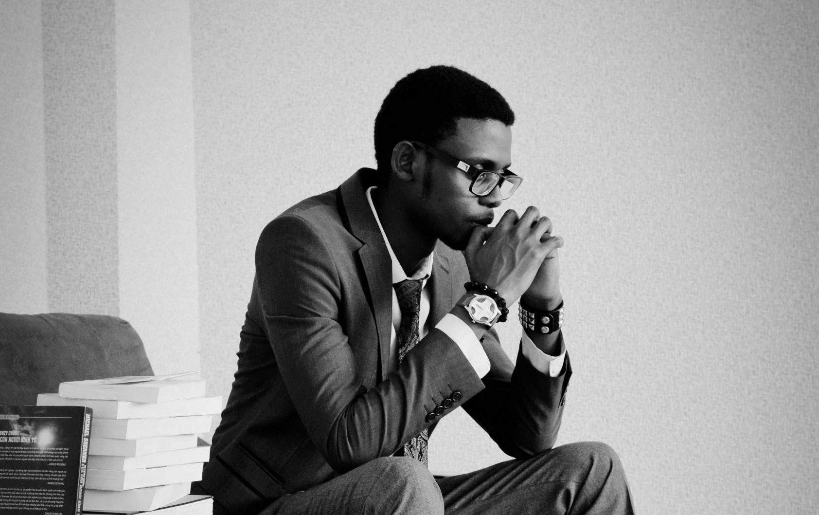 A thoughtful man in formal wear sits indoors, surrounded by books, conveying a pensive mood.
